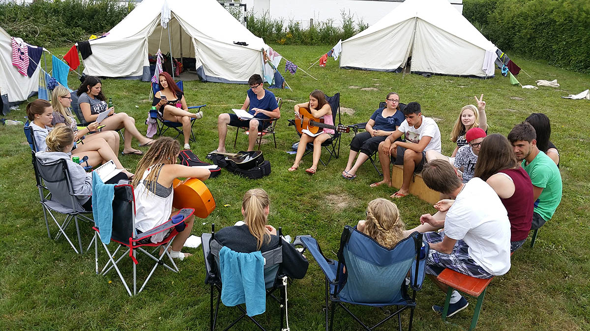 Schülergruppe sitzt im Kreis zwischen Zelten, zwei Schüler haben Gitarren in der Hand.