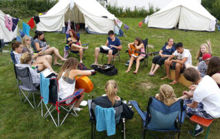 Schülergruppe sitzt im Kreis zwischen Zelten, zwei Schüler haben Gitarren in der Hand.